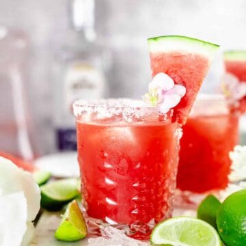 angle view shot of watermelon margarita in whisky glass garnished with purple pink flower, and watermelon slice