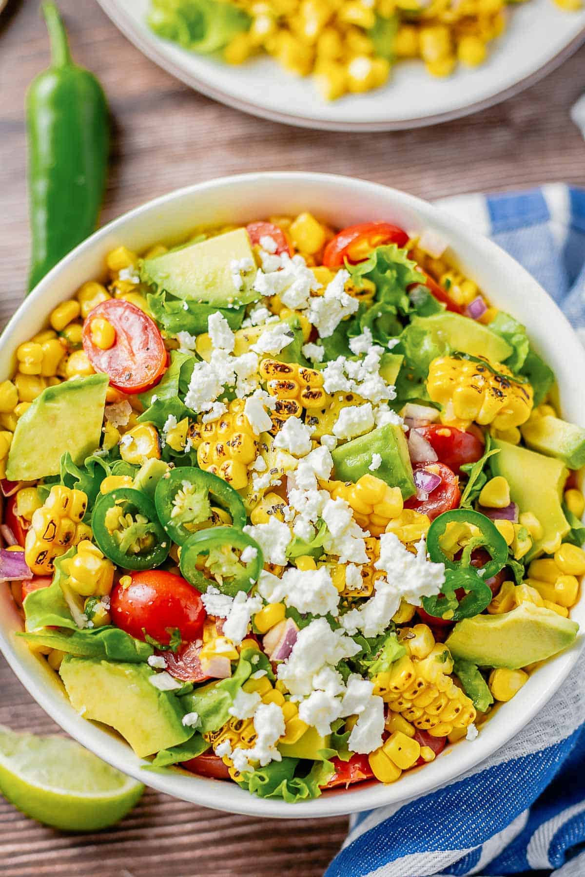 grilled corn salad with lettuce, avocado, tomatoes, onion in a white bowl on a wooden table with blue white striped towel next to it