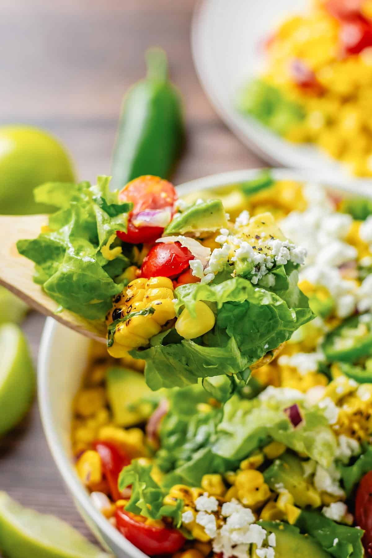 close up shot of wooden spoon with some grilled corn salad with lettuce, avocado, tomatoes, onion in a white bowl on a wooden table