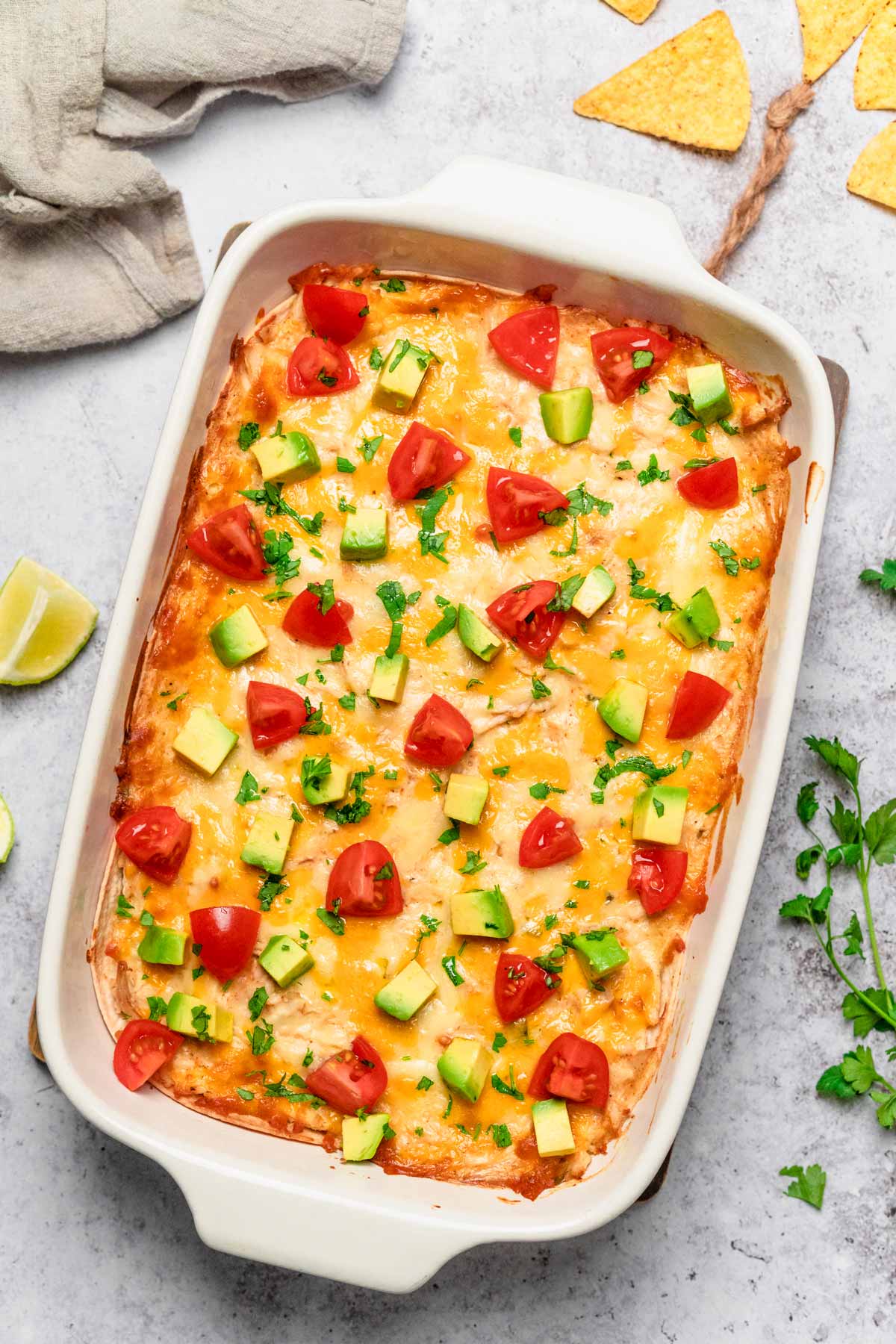 side angle shot of a taking a piece out of chicken enchilada casserole in a white baking dish with avocado and tomatoes 