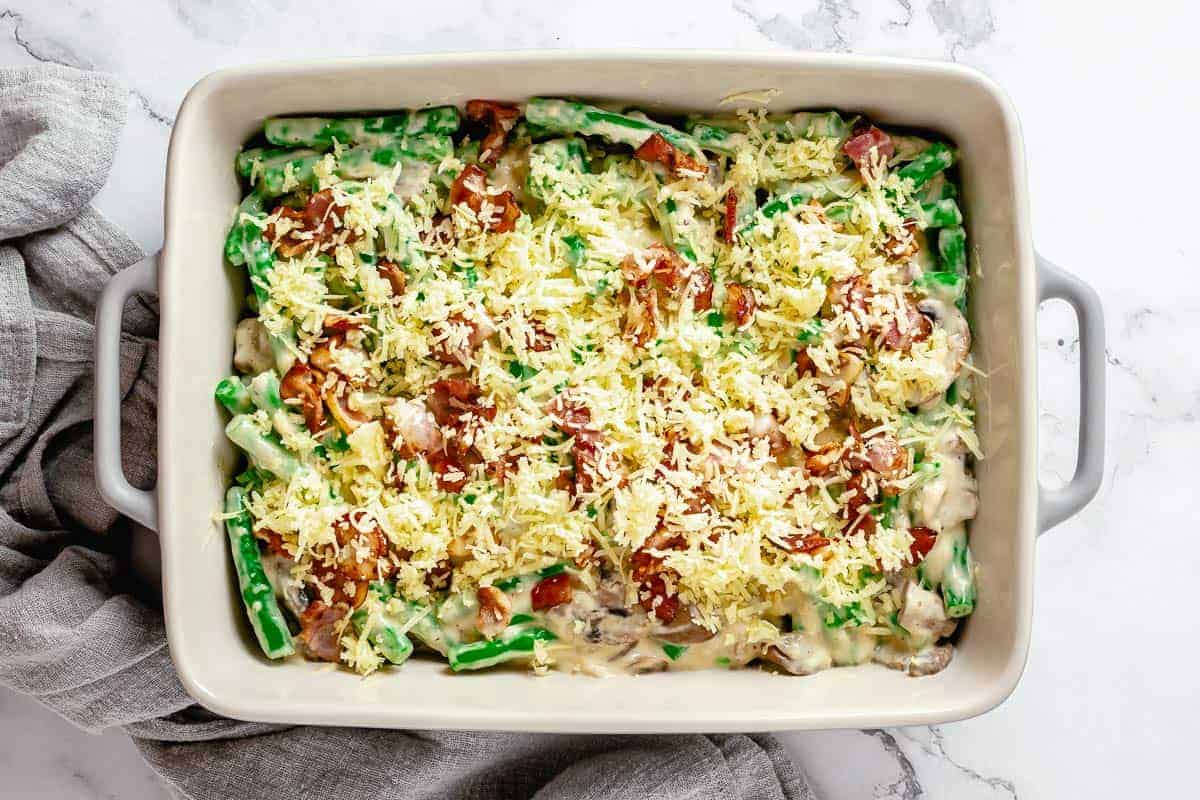 Creamy mushroom sauce with green beans in a baking dish topped with crumbled bacon and grated cheddar cheese.