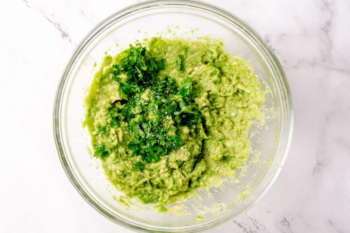 Simple guacamole in a white bowl with nacho chips around on a white marble background