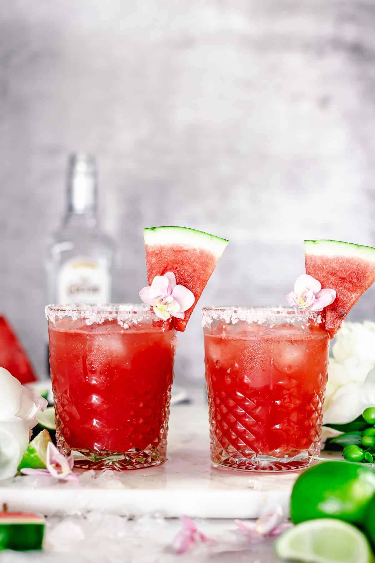 angle view shot of two watermelon margaritas side by side in whiskey glass garnished with purple pink flower, and watermelon slice